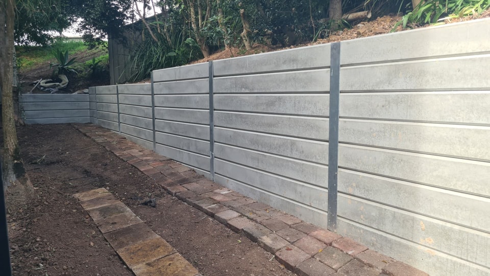 Tall grey concrete sleeper retaining wall with galvanised steel posts beside a brick-paved path in Stanwell Park